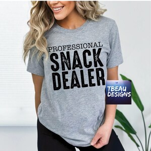 May include: A gray t-shirt with the words "PROFESSIONAL SNACK DEALER" printed in large, distressed black letters. The shirt also features a small logo that says "TBEAU DESIGNS".