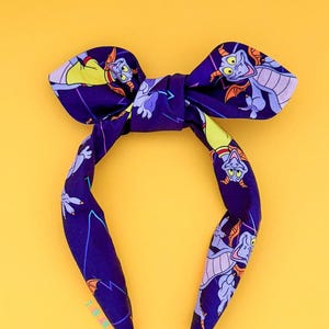 May include: A purple headband with a bow, featuring a repeating pattern of a cartoon dragon character. The dragon is in shades of purple, yellow, and orange. The background is a bright yellow.