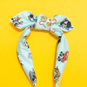 May include: A light blue headband with a bow, featuring a repeating pattern of cartoon characters. The headband is made of a silky material and is set against a bright yellow background.