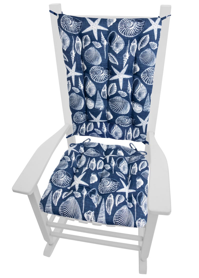 Op de afbeelding: Een witte houten schommelstoel met marineblauwe kussens. De kussens hebben een wit patroon van schelpen en zeesterren. De stoel is ontworpen voor gebruik buitenshuis en biedt comfortabele zitplaatsen voor een veranda of terras.