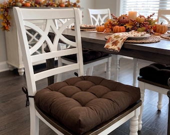 May include: A square, tufted dark brown chair cushion with fabric ties attached to the back corners. The cushion is placed on a white wooden chair with a decorative X-pattern backrest, featuring a soft, padded surface with multiple indentations.