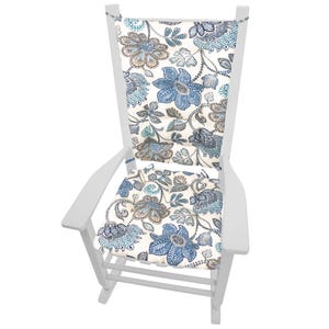 Op de afbeelding: Witte houten schommelstoel met een bloemvormig kussen. Het kussen heeft een cr&egrave;mekleurige achtergrond met blauwe, grijze en bruine bloemmotieven. De stoel heeft een hoge rugleuning en armleuningen, geschikt voor buiten of binnen.