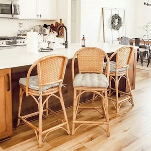 Pu&ograve; includere: Tre sgabelli da bar in rattan con cuscini a righe in una cucina. Gli sgabelli hanno schienali intrecciati e braccioli curvi. I cuscini presentano un motivo a righe blu chiaro e bianco. La cucina ha un piano di lavoro bianco e armadi in legno.