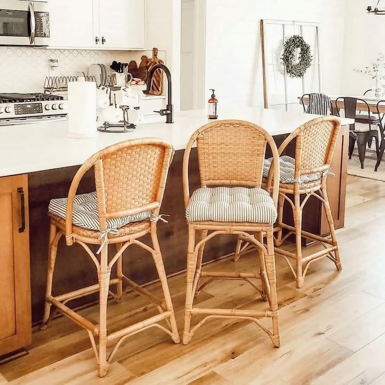 Pu&ograve; includere: Tre sgabelli da bar in rattan con cuscini a righe in una cucina. Gli sgabelli hanno schienali intrecciati e braccioli curvi. I cuscini presentano un motivo a righe blu chiaro e bianco. La cucina ha un piano di lavoro bianco e armadi in legno.