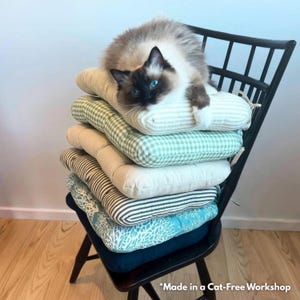 May include: A stack of five square seat cushions with various patterns including green gingham, white, brown stripes, and a teal coral print, resting on a black wooden chair. A fluffy cat with blue eyes sits on top of the cushions. Text reads: Made in a Cat-Free Workshop.