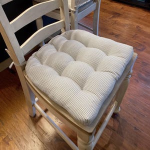 May include: A square chair cushion featuring a light gray and white gingham check pattern. The cushion is tufted with multiple circular indentations across its surface and includes fabric ties on the corners for securing it to a chair frame.