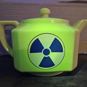 May include: A lime green ceramic teapot with a hexagonal body, a matching lid, and a handle on the left side. The front of the teapot features a prominent dark blue radiation warning symbol printed in the center of the flat hexagonal panel.