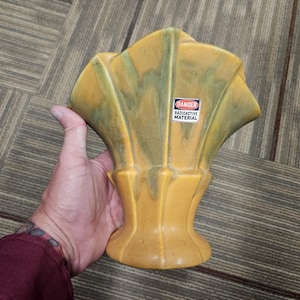 May include: A fan-shaped ceramic vase with a yellow and green glaze finish. The vase features a prominent rectangular sticker on the front with a red border and the text DANGER RADIOACTIVE MATERIAL in black and white lettering.