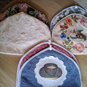 May include: A collection of decorative tea cozies in various designs. One is cream-colored with lace detailing, another features floral embroidery, and a third has a blue paisley pattern with a teacup illustration. The cozies are arranged on a wooden surface.