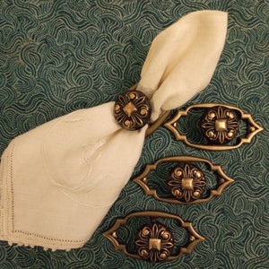 May include: Four ornate gold napkin rings with a decorative floral design. The rings are made of metal and have a dark brown finish. One ring is holding a white linen napkin.