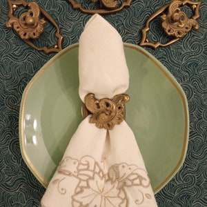 May include: A white linen napkin with an embroidered floral design is held by a gold napkin ring with a decorative floral design. The napkin ring is on a light green plate with a gold rim. The plate is on a dark green patterned surface.