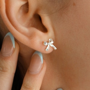 May include: A close-up of a person's ear with a small silver bow earring. The person has light brown hair and is wearing a light blue shirt.