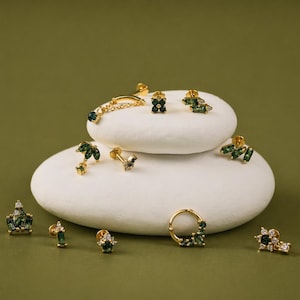 Puede incluir: Una colección de pendientes dorados con detalles de gemas verdes y transparentes. Los pendientes se exhiben sobre piedras blancas de forma ovalada sobre un fondo verde oliva liso. Los estilos incluyen tachuelas, aros y diseños colgantes.