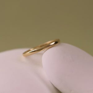 以下が含まれることがあります： 滑らかな有機的なデザインのゴールドリングが、柔らかい緑色の背景に対して、白く丸みを帯びた表面に置かれています。 リングの磨かれた表面は光を反射し、その湾曲した形状を強調しています。 ジュエリー。