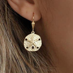 Peut inclure: Boucles d'oreilles en forme de dollar des sables de couleur or. Les boucles d'oreilles présentent un motif détaillé de dollar des sables avec une étoile de mer au centre et de petits trous sur les bords. Les boucles d'oreilles sont suspendues à un crochet doré.