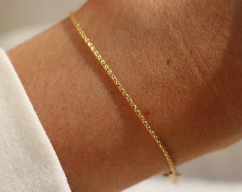 Puede incluir: Una delicada pulsera de cadena de oro con un diseño de eslabones finos tipo espiga. La pulsera se lleva alrededor de la muñeca, mostrando un acabado metálico pulido que refleja la luz a lo largo de su estructura delgada, continua y trenzada.