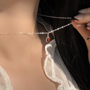 Könnte beinhalten: Eine zarte silberne Kettenkette mit einem kleinen, quadratischen Anhänger mit einem roten Edelstein.