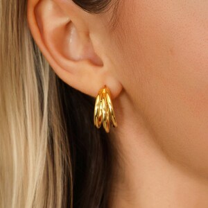 May include: Gold triple hoop earrings. The earrings are made of a shiny, reflective metal and are worn on the ear. The hoops are close together, creating a layered effect. The earrings are a simple yet elegant accessory.