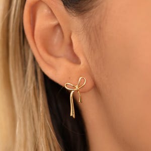May include: A close-up of a gold-coloured bow-shaped stud earring. The earring is worn on a person's ear. The bow design has a looped top and two dangling ends. The person has blonde hair.