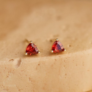 May include: A pair of gold stud earrings, each featuring a triangular, vibrant red gemstone. The earrings are set on a textured, light brown surface, with a soft yellow background. The gemstones have a faceted appearance, catching the light.