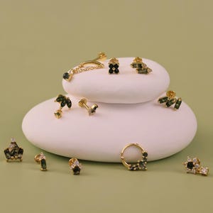 May include: A collection of gold earrings with dark green and clear gemstones are arranged on white, rounded platforms against a pale green background. The earrings showcase diverse designs, including studs, hoops, and drop styles.