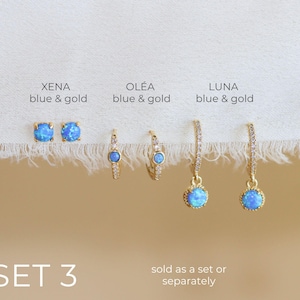 May include: A selection of gold-coloured earrings featuring blue opal stones. The earrings include square studs, small hoops, and drop styles. The image text reads "XENA blue & gold", "OLÉA blue & gold", and "LUNA blue & gold". The text also reads "SET 3" and "sold as a set or separately".