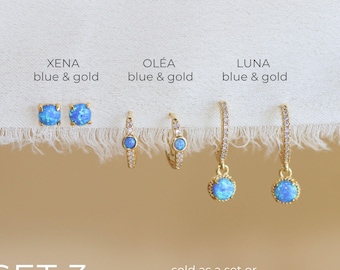 May include: Three pairs of gold-tone earrings labeled XENA, OLÉA, and LUNA, all featuring blue stones. The XENA pair are studs, the OLÉA pair are small hoops, and the LUNA pair are hoop earrings with dangling blue stone charms. Text reads SET 3.