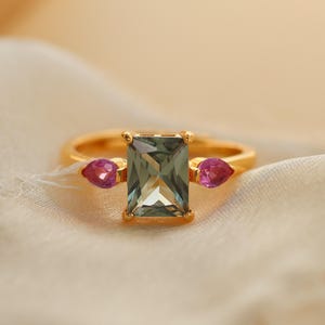 May include: A gold ring featuring a rectangular, light green gemstone as the centerpiece, flanked by two smaller, teardrop-shaped pink gemstones. The ring is set against a soft, cream-colored fabric.