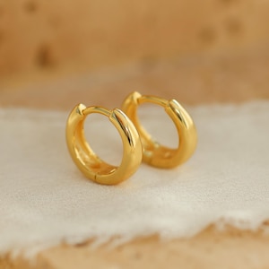 May include: A pair of small, gold-colored hoop earrings. The earrings are round and have a smooth, polished surface. They are displayed on a white, textured surface against a blurred, light brown background.