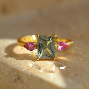 May include: A gold ring featuring a rectangular, light green gemstone as the centerpiece, flanked by two smaller, pink teardrop-shaped stones. The ring is set on a neutral-toned surface, showcasing the jewelry's design and color contrast.