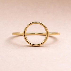 May include: A gold ring featuring a thin, polished band that connects to a centred, open circular element. The metal has a smooth, reflective surface, and the design consists of a simple, continuous loop shape integrated into the band structure.