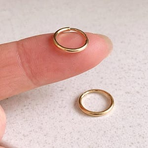 May include: Two small, gold-coloured hoop earrings. One earring is on a fingertip, and the other is on a white surface. The earrings are circular and appear to be made of metal. The background is blurred, with a focus on the earrings.
