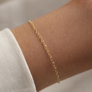 May include: A delicate gold chain bracelet featuring small, uniform oval-shaped links. The bracelet is worn around the wrist, showcasing a simple and refined design with a polished metallic finish that catches the light across its surface.