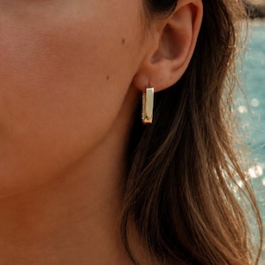 May include: A gold hoop earring with a rectangular, flat-edged design worn in the earlobe, paired with a delicate gold chain necklace featuring a small, textured pendant that rests against the collarbone area.
