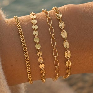May include: A set of four gold-toned bracelets worn on the wrist. The collection includes a classic curb chain, a delicate disc chain, an elongated oval link chain, and a textured oval disc chain, all featuring a polished metallic finish.