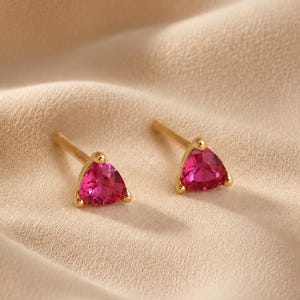 May include: A pair of stud earrings featuring triangular-cut vibrant pink gemstones set in gold metal. Each earring has a post back and a polished gold setting that secures the faceted pink stone, creating a bright and reflective appearance.