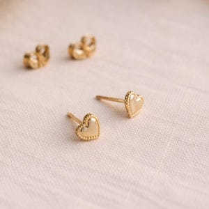 Può includere: Orecchini a bottone a forma di cuore color oro con bordo perlato. Gli orecchini sono esposti con due chiusure abbinate su una superficie chiara. Gioielli.