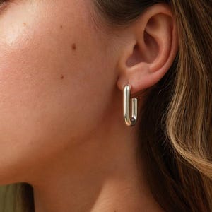 May include: Silver rectangular hoop earrings. The earrings have a polished finish and a modern, minimalist design. The image shows a close-up of the earrings on an ear, highlighting their shape and shine. The earrings are a simple yet elegant accessory.
