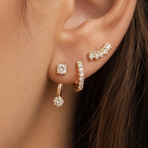 May include: A set of gold earrings featuring a small stud with a round clear stone, a gold hoop earring with a dangling round clear stone, a small gold hoop with multiple clear stones, and a curved gold ear climber with five clear stones.