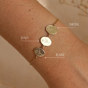 May include: A gold bracelet featuring three oval pendants with floral engravings. The pendants are inscribed with "MOM", "DAD", and "BABY". The bracelet is shown on an arm.