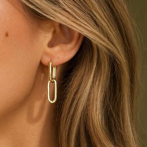 May include: Gold-coloured, linked hoop earrings. The earrings feature a small, circular hoop that connects to a larger, elongated oval shape. The earrings are shown on a person with blonde hair.