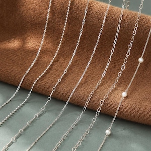 May include: A collection of six distinct silver-toned metal chains displayed side by side. The styles include a beaded chain, a bar link chain, a cable chain, a heart-shaped link chain, and a delicate chain accented with small white round pearls.