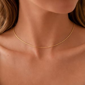Peut inclure: Un collier chaîne fine de couleur dorée à maillons carrés, reposant sur le cou. Le bijou présente un design uniforme et continu, créant une texture métallique lisse. La chaîne est simple, délicate et se positionne à la base de la gorge.