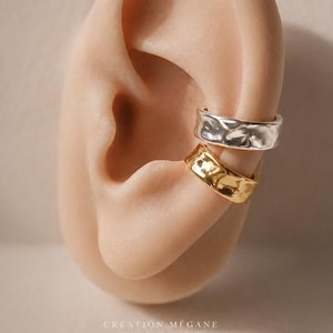 May include: Two ear cuffs, one silver and one gold, are displayed on an ear model. The silver cuff is on top, and the gold cuff is below. Both cuffs have a hammered texture. The background is a neutral colour.