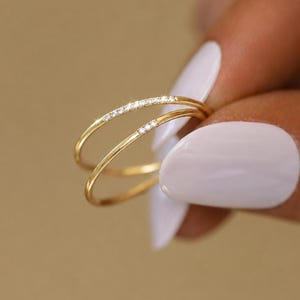 May include: Two delicate gold rings, one with a row of small, clear gemstones. The rings are held against a soft, neutral background. The rings are a jewelry item.