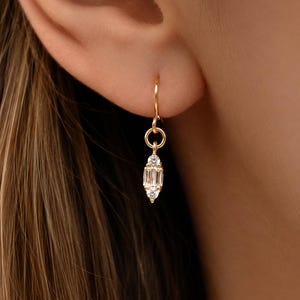 May include: A gold drop earring featuring a small circular hoop connected to a vertical pendant. The pendant consists of a central baguette-cut clear gemstone flanked by two smaller round-cut clear gemstones, all set in a polished gold metal frame.