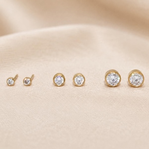 Peut inclure: Trois paires de boucles d'oreilles à clous en or, chacune dotée d'une pierre ronde transparente sertie clos. Les boucles d'oreilles sont disposées en rangée de la plus petite à la plus grande, avec une finition en or poli et des pierres facettées.