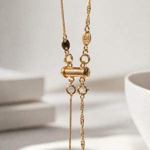 Può includere: Una collana dorata con chiusura cilindrica e sottili maglie a catena. La collana presenta un elemento centrale a forma di barilotto ed è posta su uno sfondo morbido e neutro. Il design è semplice ed elegante.