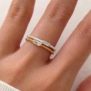 Peut inclure: Deux bagues délicates, une dorée et une argentée, chacune ornée d'une petite pierre précieuse rectangulaire et transparente. La bague dorée est empilée sous la bague argentée. Les anneaux ont un design torsadé texturé.