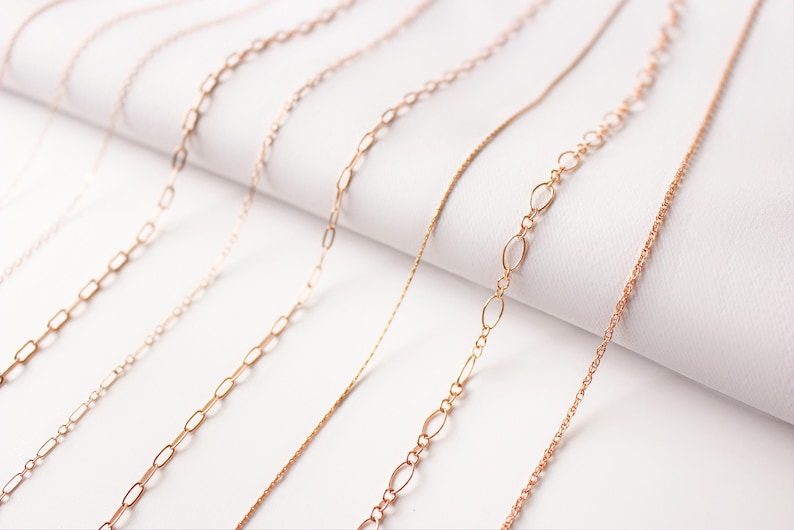 May include: A collection of seven gold chains of different styles and thicknesses, laid out on a white surface.